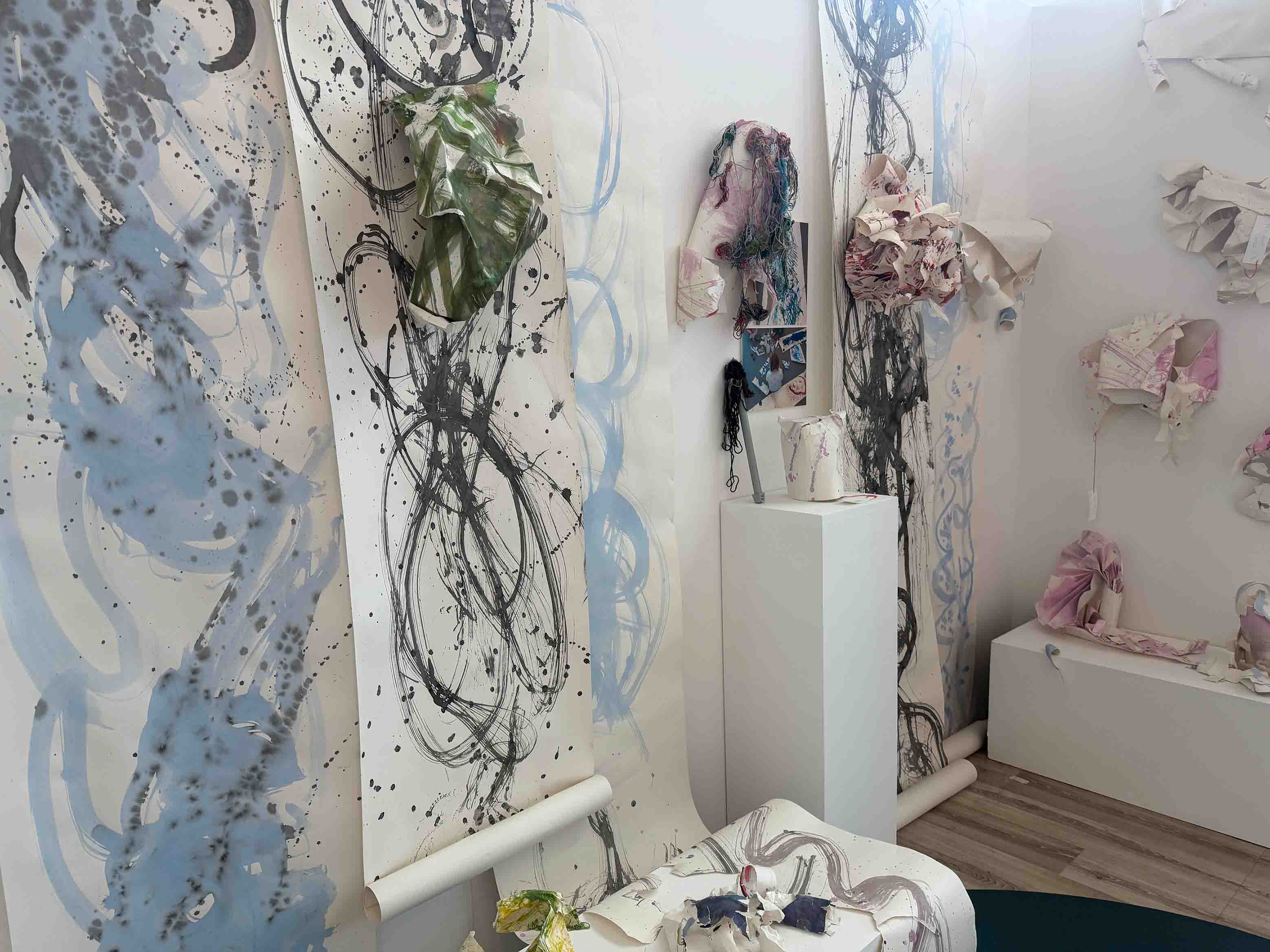 Long pieces of paper covered in painted marks draped down the wall with small paper sculptures on plinths