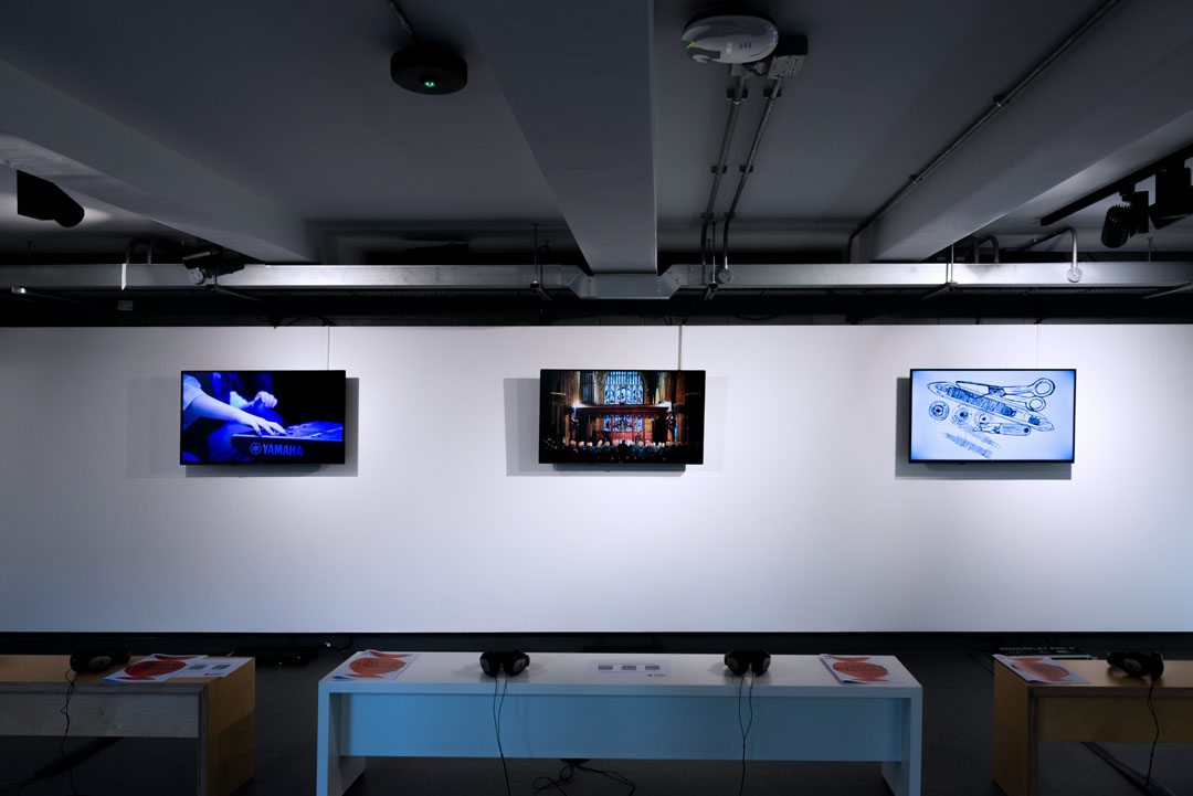 Three TV's on a white wall with a white bench in front of them with headphones on.