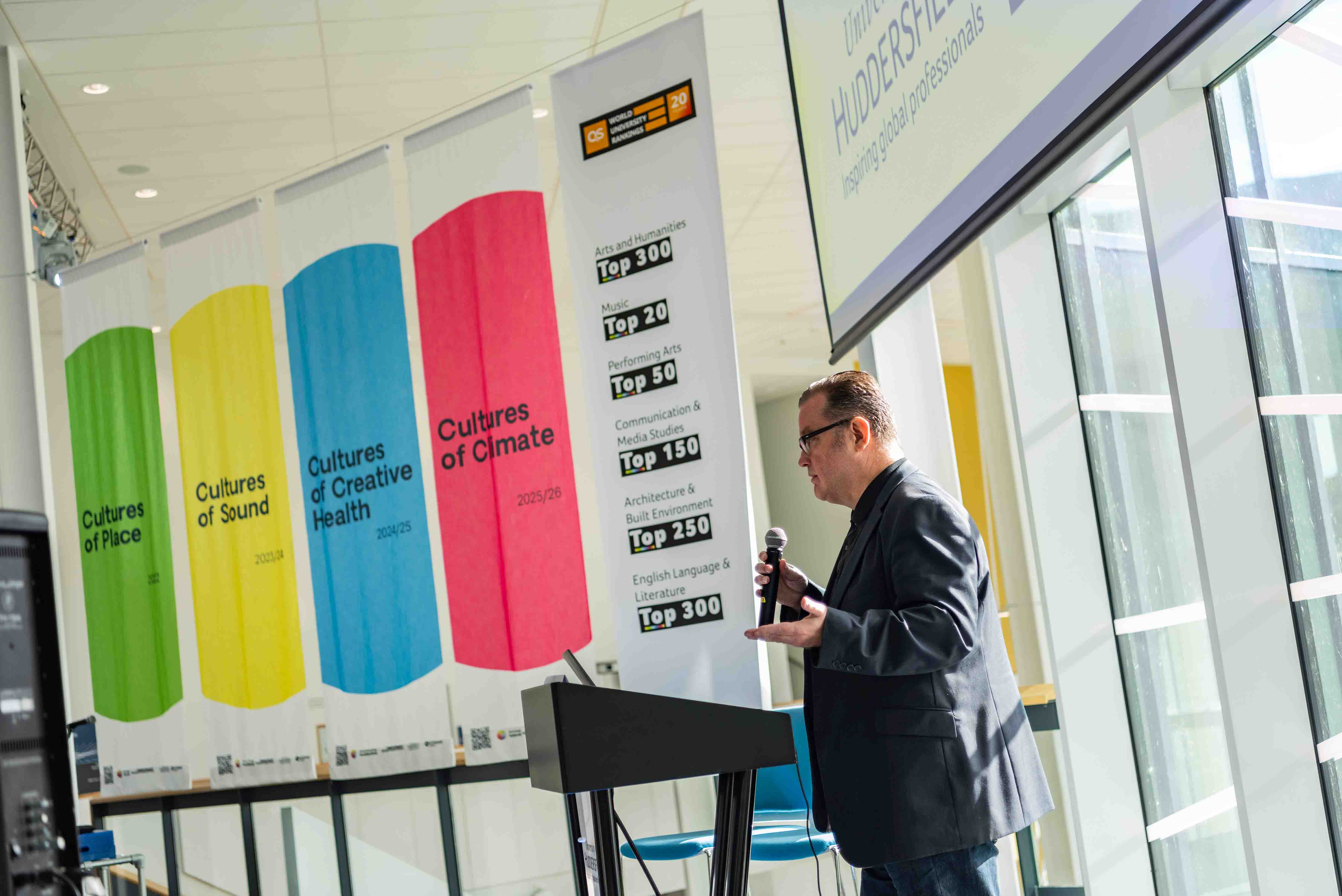 A man in a suit talking into a microphone with 4 colourful banners in the background