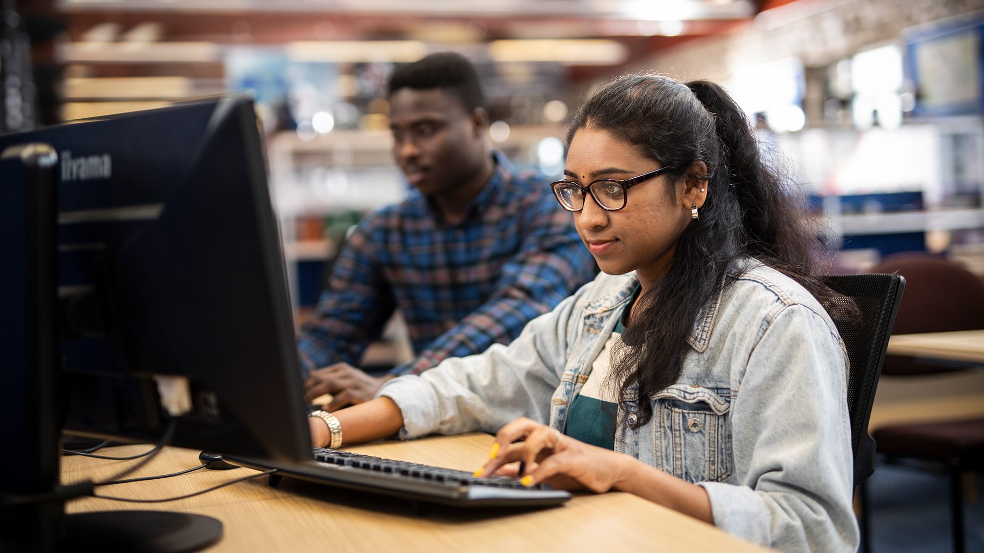 A student working on a computer, with another student in distance also working on one.
